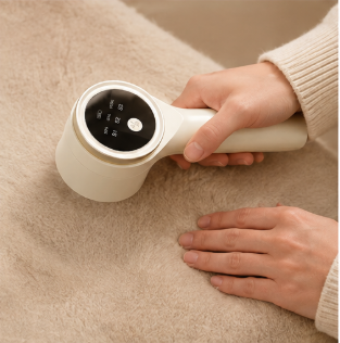 A handheld white and beige fabric shaver or lint remover being used on a beige textured blanket, with a person's hands visible holding and demonstrating it.