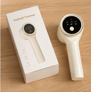 A handheld hairball trimmer shown next to its white retail box on a light wood surface. The device is cream-colored with a black circular display screen and a central white button.